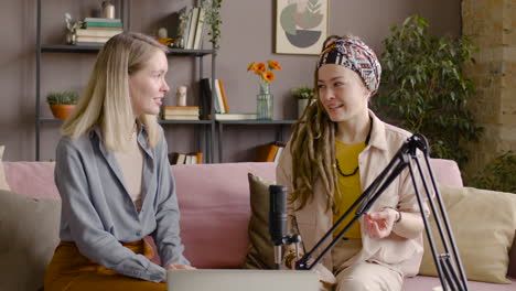 two women recording a podcast talking into a microphone sitting on sofa in front of table with laptop