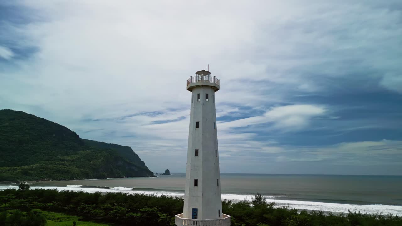 Un filmato aereo realizzato con drone riprende un alto faro bianco che si erge su un paesaggio costiero verde, con colline boscose sullo sfondo e onde oceaniche che si infrangono su una tranquilla costa tropicale sotto nuvole in movimento.