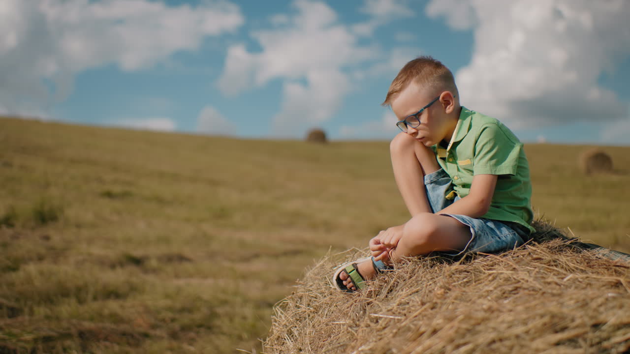 маленький мальчик сидит на пучке сена с наклоненной головой, потерянный в мыслях, окруженный огромным открытым полем под ярким голубым небом, захватывая тихий, мирный момент в сельской местности в теплый солнечный день.