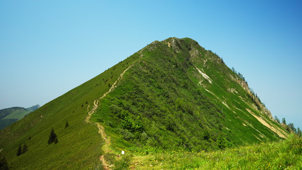 A stunning ridgeline trail near Golica, Slovenia, with steep grassy slopes on both sides. The narrow mountain path across a green crest with forested hills and peaks stretching far into the horizon.