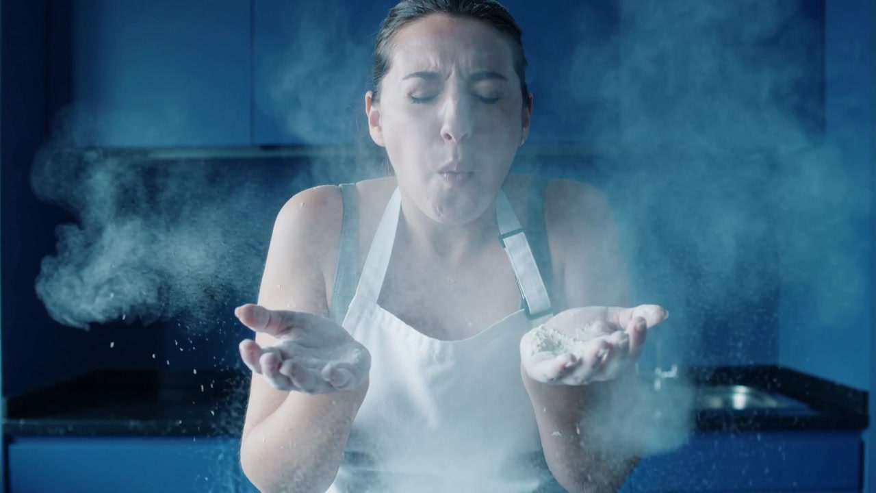 Cupping handfuls of white powder near face, woman in apron blowing cloud across kitchen for baking