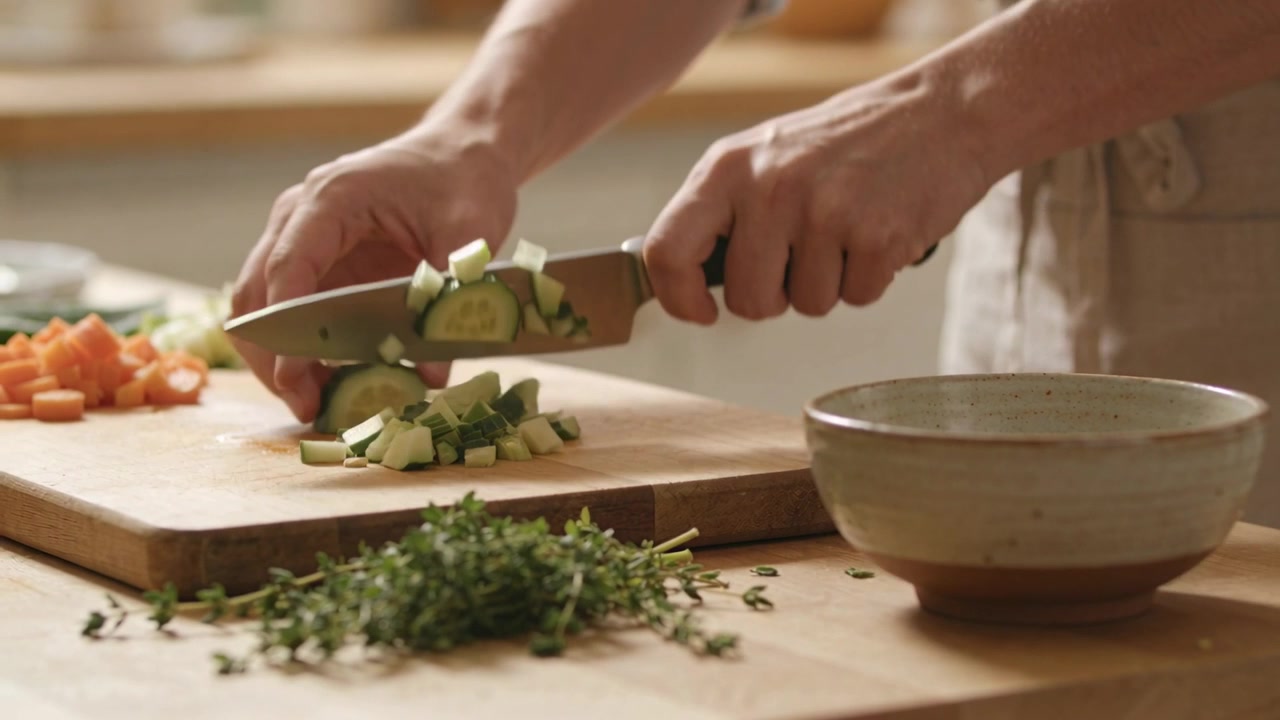 Mani che affettano, tenendo un coltello da chef, tagliano un cetriolo a cubetti su un tagliere di legno con carote, erbe aromatiche e una ciotola.