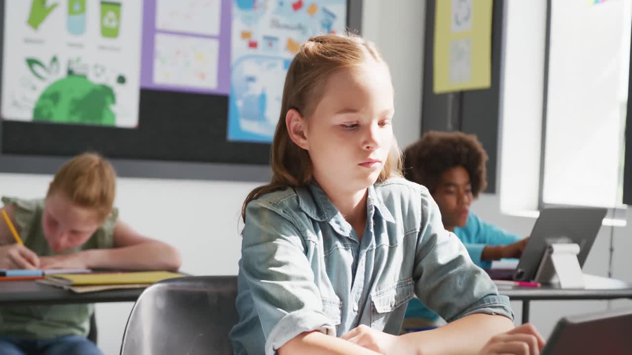 Video di una scolara caucasica concentrata alla scrivania che usa un tablet in una classe eterogenea, spazio per il testo.