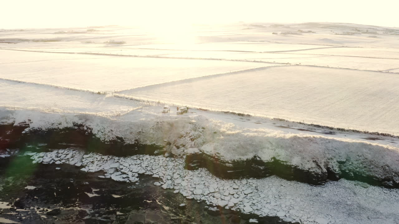 Drone descends as sun flare spreads light across surfers hanging out at top of sea cliff by cars on cold snowy winter day