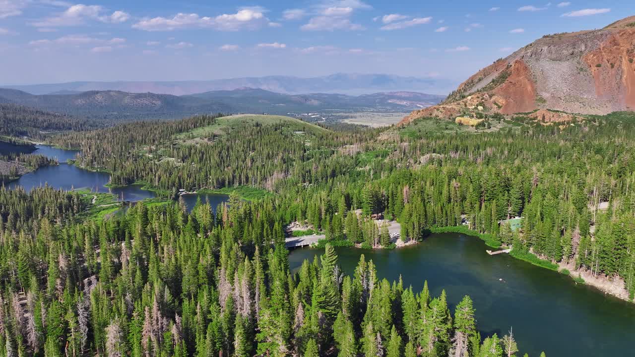 Drone pans across Lake Mary the Mammoth Lakes Basin revealing blue alpine water dense pine forest and red volcanic cliffs with Eastern Sierra peaks beyond. Bright summer daylight smooth lateral motion