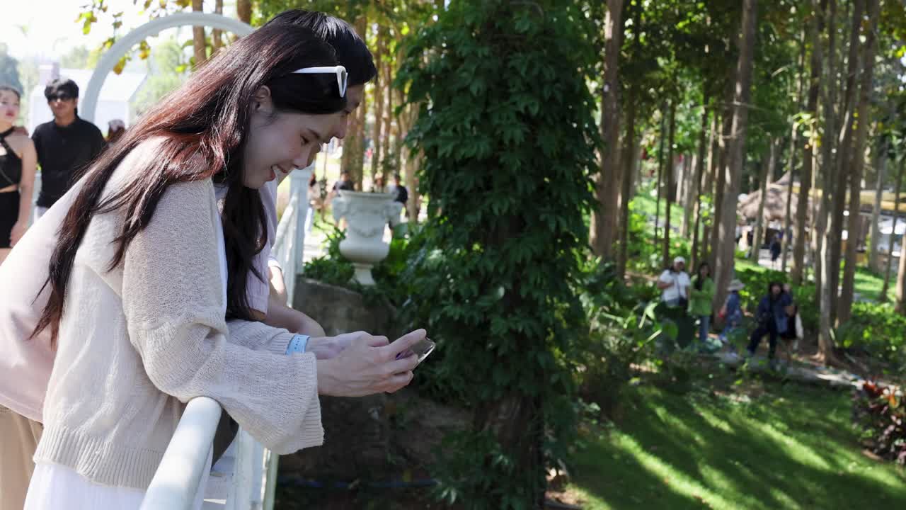Giovane donna sorridente si appoggia a una ringhiera bianca di un ponte, usando lo smartphone in un luminoso parco forestale