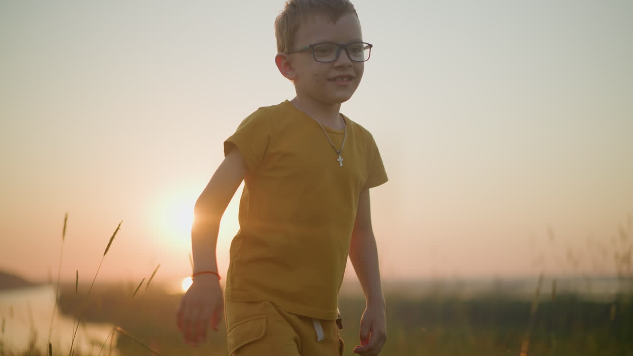 노란색 셔츠와 쇼트를 입은 어린 소년은 빛이 비치는 풀에서 기게 점프합니다. 따뜻한 빛은 어나와 그를 둘러싼 아름다운 빛을 만니다.