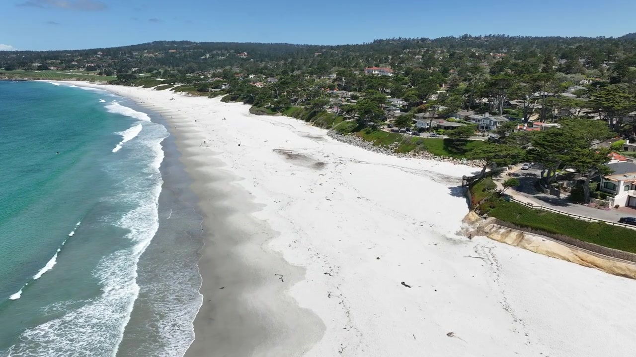 Wide cinematic aerial drone footage of Carmel-by-the-Sea, California, showcasing expansive white sand beaches, crystal-clear turquoise Pacific Ocean water, rolling ocean waves, luxury coastal homes