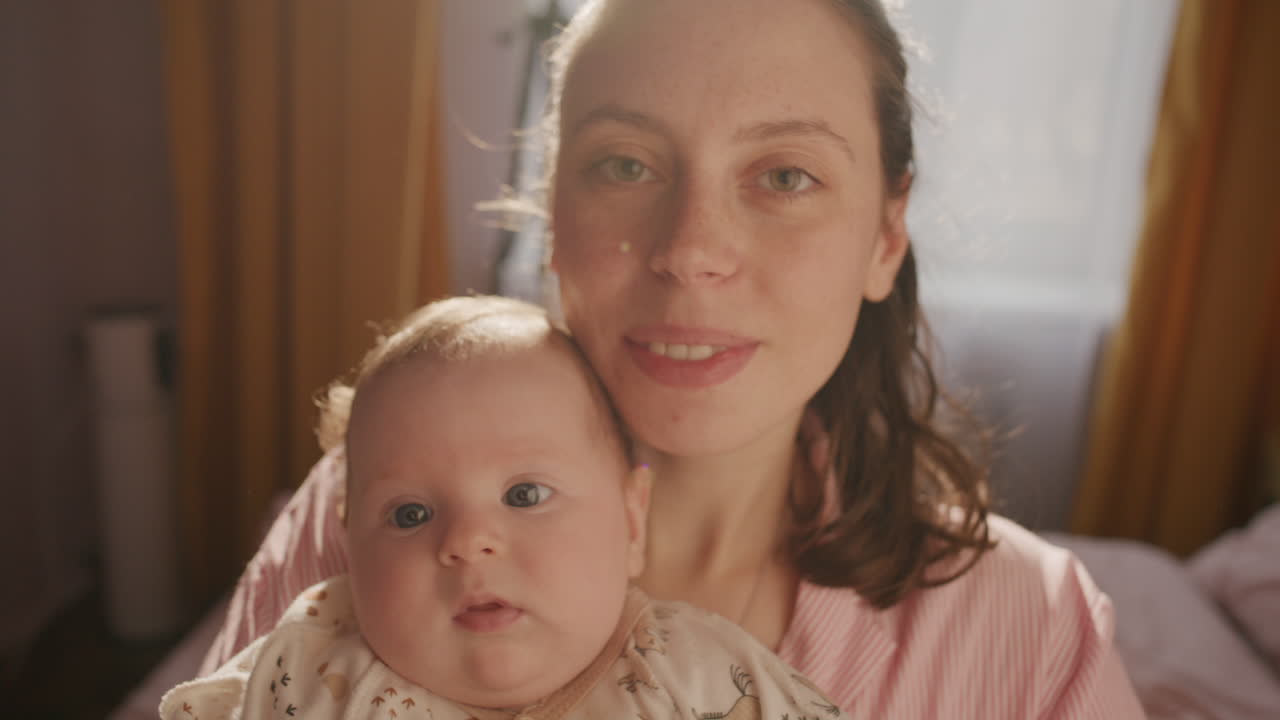 Porträt einer jungen, lächelnden Mutter und eines Babys, die zusammen in einem sonnendurchfluteten Raum posieren