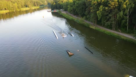 aerial tracking shot of active wake boarder surfing on natural pond surrounded by forest trees in wilderness