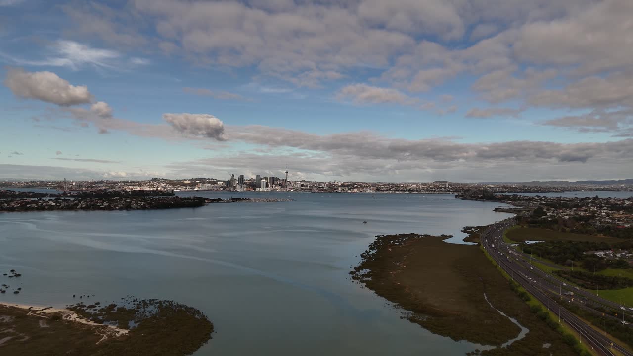A scenic 4K 60fps aerial flight over the tranquil waters of Shoal Bay, revealing the stunning Auckland CBD skyline, including the Sky Tower, in the background. A perfect shot of NZ city life