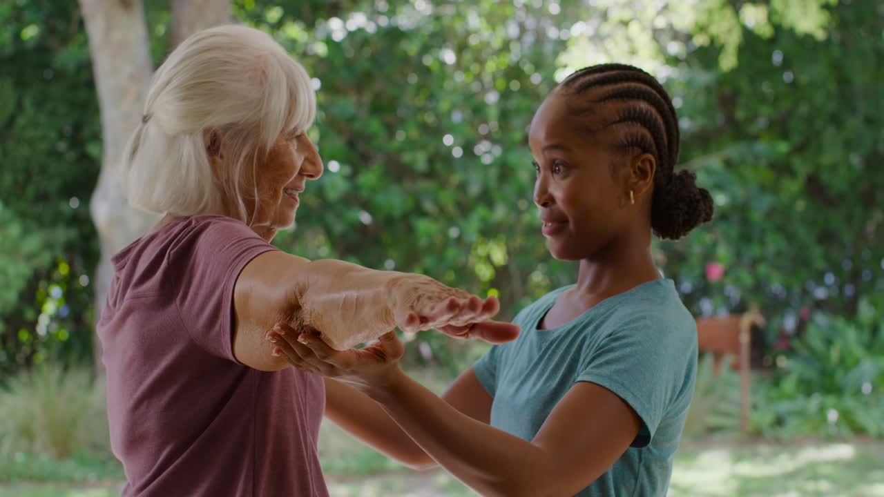 Mujer mayor sujetando los brazos de una mujer afroamericana que le ayuda a mantener el equilibrio en un patio, apoyando sus manos bajo los brazos de la mayor.