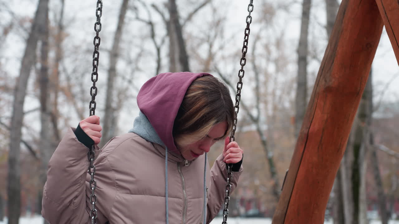 молодая леди в зимнем наряде подходит к качелю, ступает на нее и начинает качеться, покрытая снегом земля, голые деревья и городские здания создают спокойную, ностальгическую зимнюю атмосферу в открытом парке