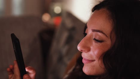 Young-Italian-woman-smiling-while-scrolling-through-a-smart-phone-while-sat-on-the-sofa-at-home