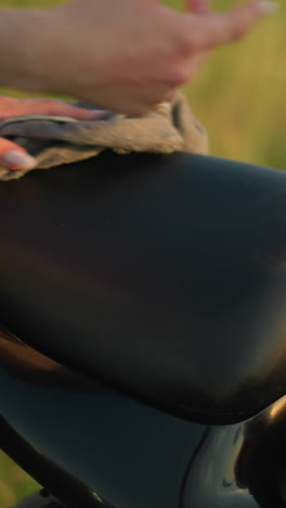 a close-up of a hand cleaning the seat of a motorcycle with a cloth in a grassy field, the person is carefully wiping down the bike