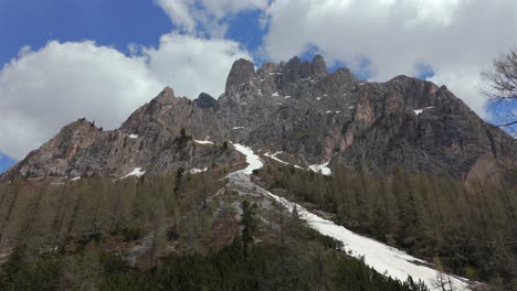 impresionante vista de un pico de montaña rocosa con manchas de nieve y pinos bajo un cielo azul brillante con nubes esponjosas