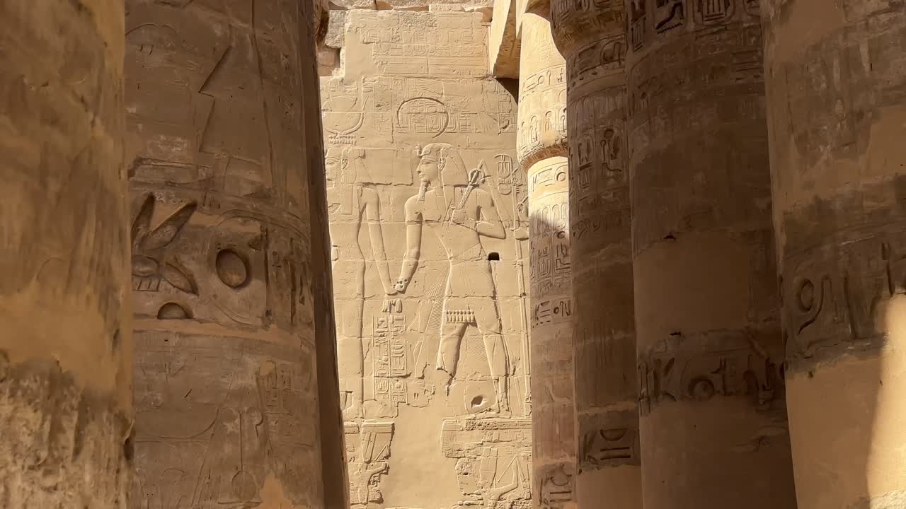 Elokuvamainen näkymä massiivisista kivipylväistä ja suuresta seinäreliefistä, jossa on muinaisia egyptiläisiä hahmoja ja hieroglyfejä Karnakin suuren pylvässalin sisällä.