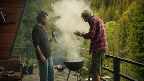 Two-guys-check-the-food-they-cook-on-the-grill-and-blow-high-fives-to-each-other,-because-they-are-doing-great.-Thick-white-smoke-comes-from-the-brazier-against-the-backdrop-of-mountains-and-coniferous-forests