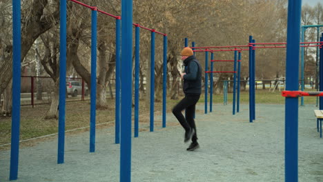 a coach performs a fast-paced exercise alone in an outdoor stadium, wearing a brown beanie and black outfit, he is surrounded by blue and red iron bars around