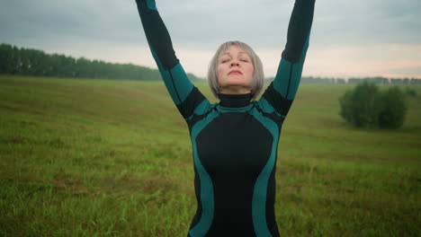 woman in green and black suit standing with head slightly lifted, eyes closed, lifting hands up in a serene motion while practicing yoga in a vast open field under a cloudy sky