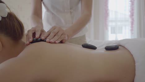 Professional-hot-stone-massage,-therapist-is-using-stones-to-massage-persons-back-while-lying-on-the-massage-table,-well-being-and-health-concept