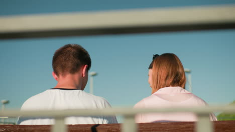 back view of a lady with glasses on her head smiling warmly at a man seated next to her on a bench, framed by an iron railing and a bright sky