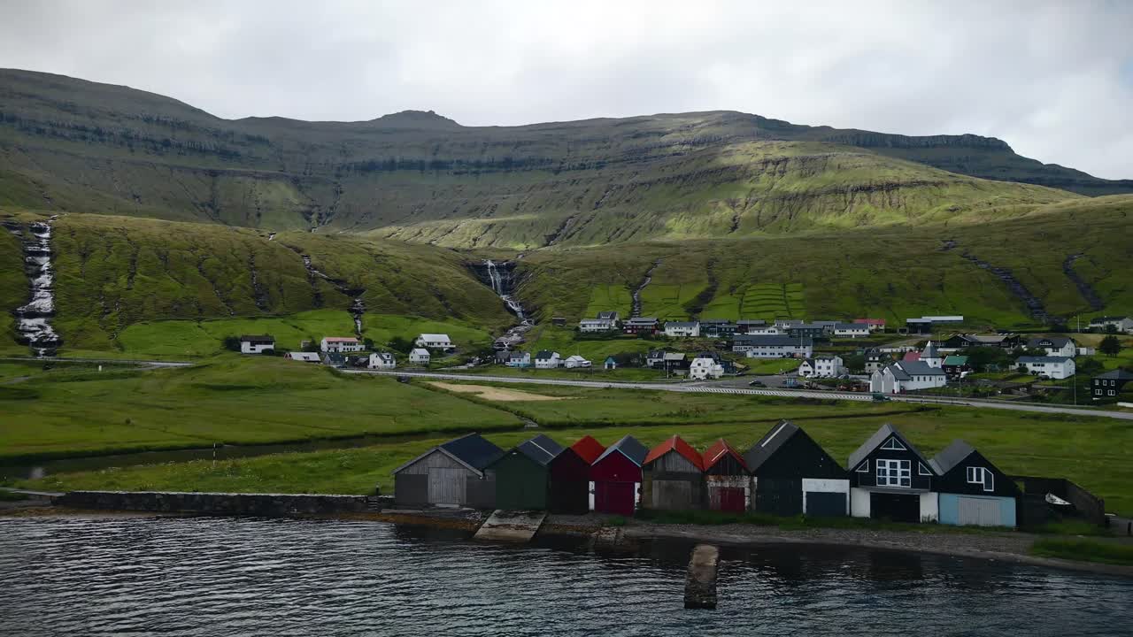 페로 제도의 스트레이모이 섬에 있는 호스비크 마을 연안에 있는 다채로운 보트 하우스는 푸른 산에서 아오르는 폭포와 함께 평온한 풍경을 보완합니다.