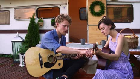 Bearded-guy-teaching-her-girlfriend-play-the-guitar-outdoors-sitting-close-to-their-trailer-house.-Holding-acoustic-guitars,-man-showing-how-to-tap-particular-chords