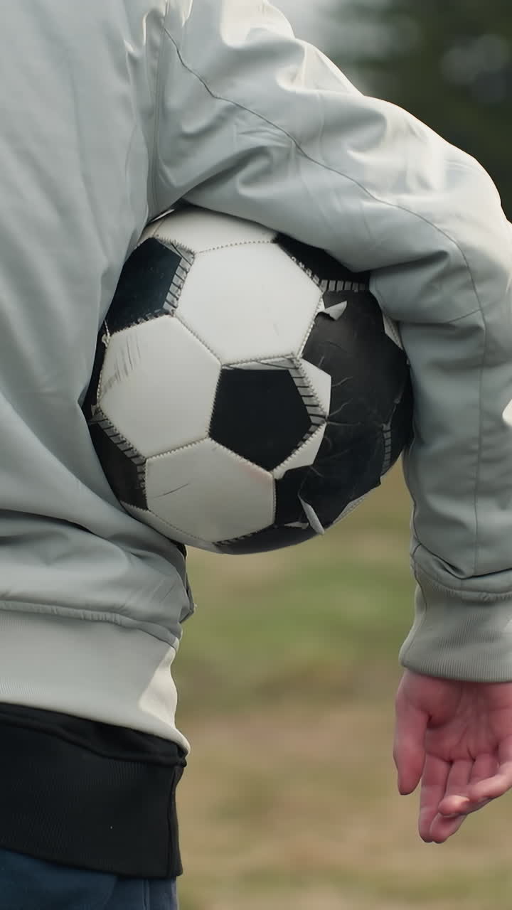 Close-up of a person standing outdoors in a gray outfit, holding a soccer ball under their right arm, with their left hand in the pocket