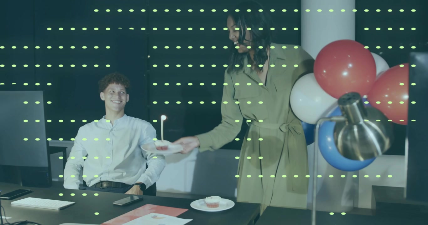 Male colleague typing in office, female peer bringing cupcakes, balloons sparking birthday surprise
