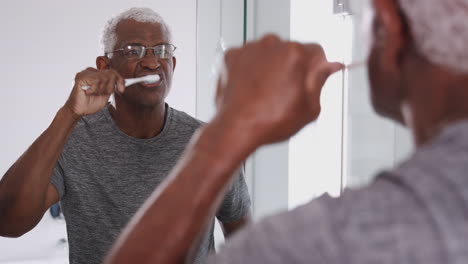 senior man wearing pajamas looking at reflection in bathroom mirror brushing teeth