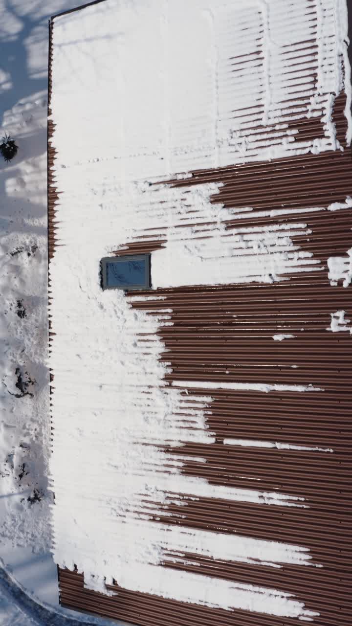 Flying above a roof covered in snow. Snow melting on a roof in a sunny winter day. Vertical aerial drone view of a brown house roof.