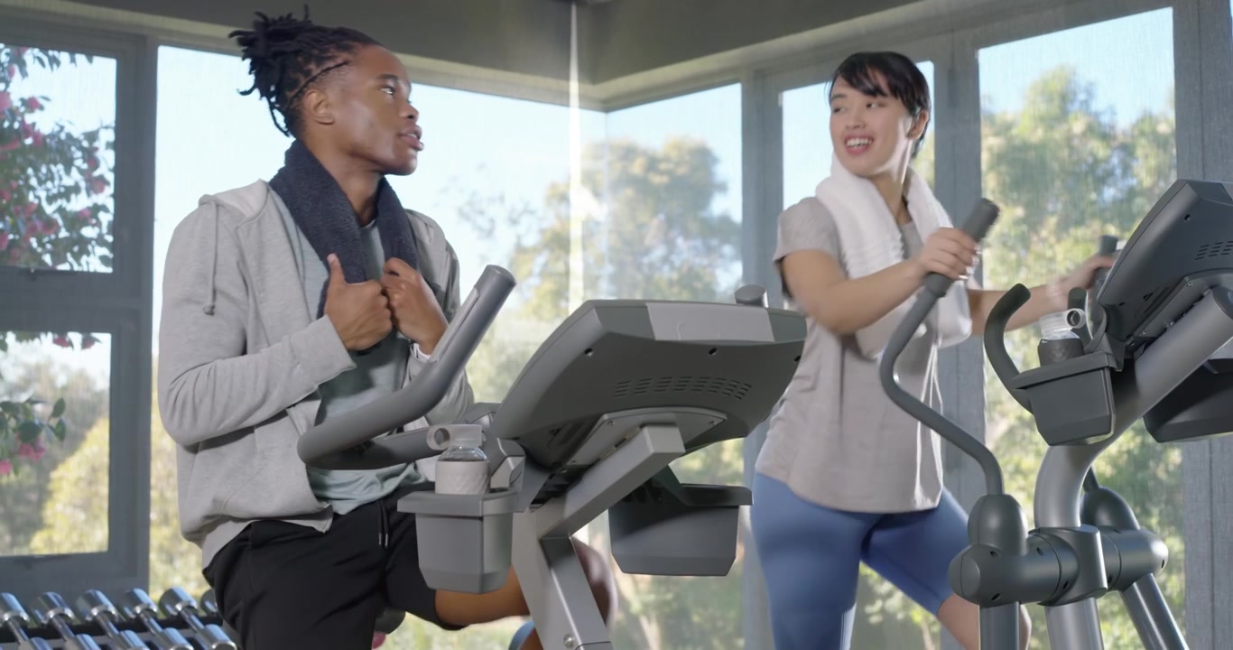 Amici diversi in palestra si riscaldano per il cardio, un uomo pedala una cyclette, una donna usa l'ellittica, asciugamani, acqua