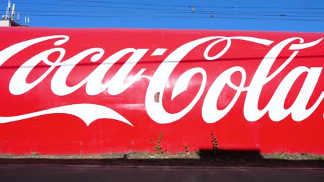 Under a bright, clear sky, a low-angle view captures pedestrians passing by a wall in Ulaanbaatar, Mongolia, featuring the Coca-Cola logo and vibrant red color, promoting the American soft drink.