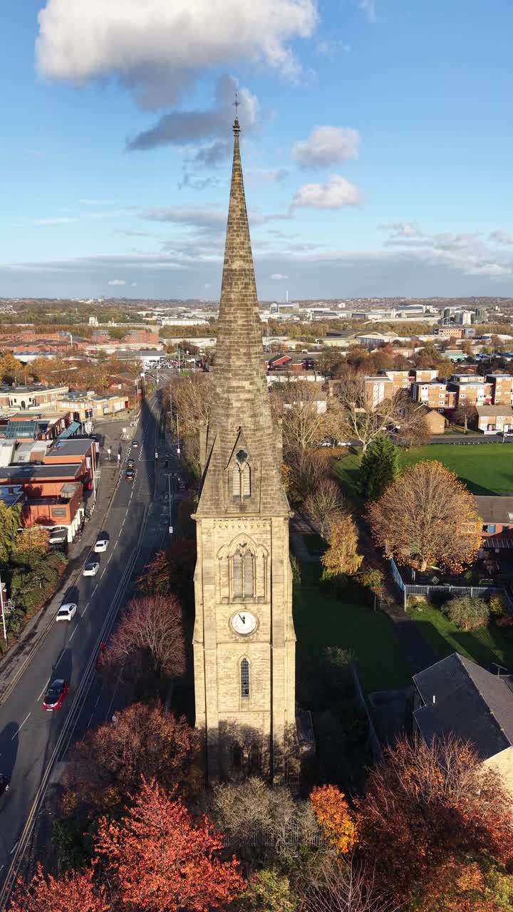 Vista con drone in stile social media verticale di una guglia di chiesa storica a Hedge End, Hampshire, che si erge sopra le strade della città, alberi autunnali, edifici residenziali e un cielo blu sereno.