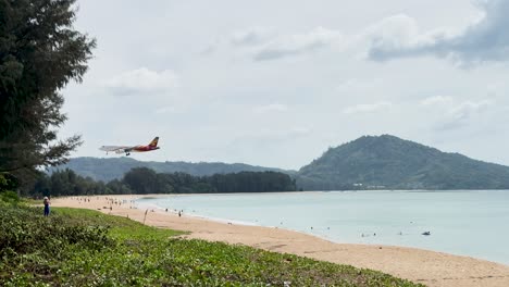 an airplane descends over mai khao beach, phuket, with a scenic backdrop of mountains and ocean under a cloudy sky