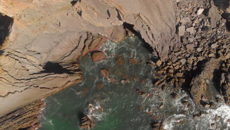 top-down shot of waves crashing at the bottom of the rocky cliff on the shore in costa da morte, galicia, spain - aerial