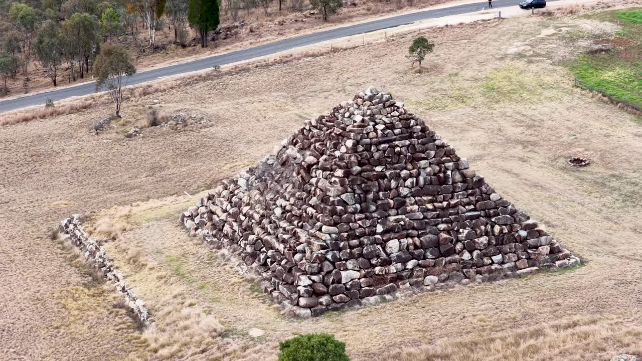 Drone circles a large stone pyramid structure in a dry, grassy field under daylight, revealing changing perspectives and surrounding rural landscape