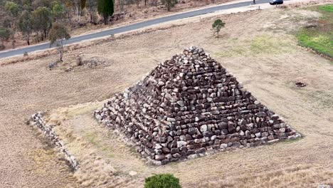 drone circles a large stone pyramid structure in a dry, grassy field under daylight, revealing changing perspectives and surrounding rural landscape