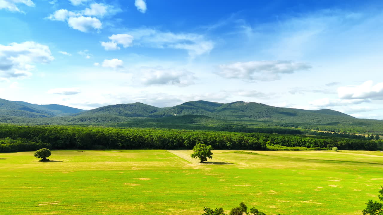 Vast green landscape, mountains ahead. Lush green field stretches below mountains under a bright blue sky with fluffy clouds