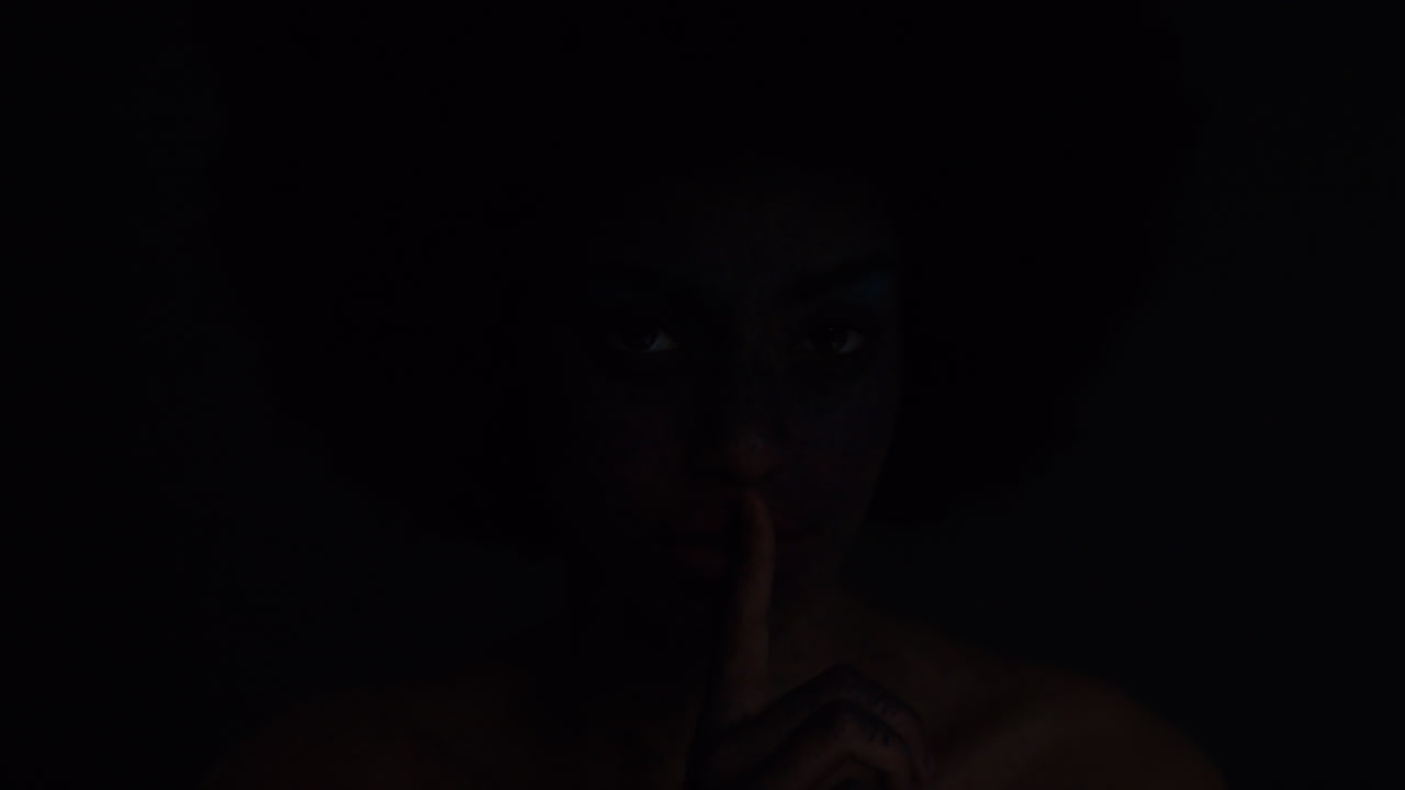 retrato de una hermosa mujer afroamericana con cara pintada arte corporal que muestra silencio gesto femenina misteriosa que mantiene la luz secreta parpadeando en el fondo oscuro concepto de expresión creativa