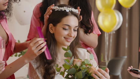 grupo de amigas que componen a la novia y le ponen una banda para el cabello sosteniendo un ramo sentadas en la cama en una reunión nupcial 1