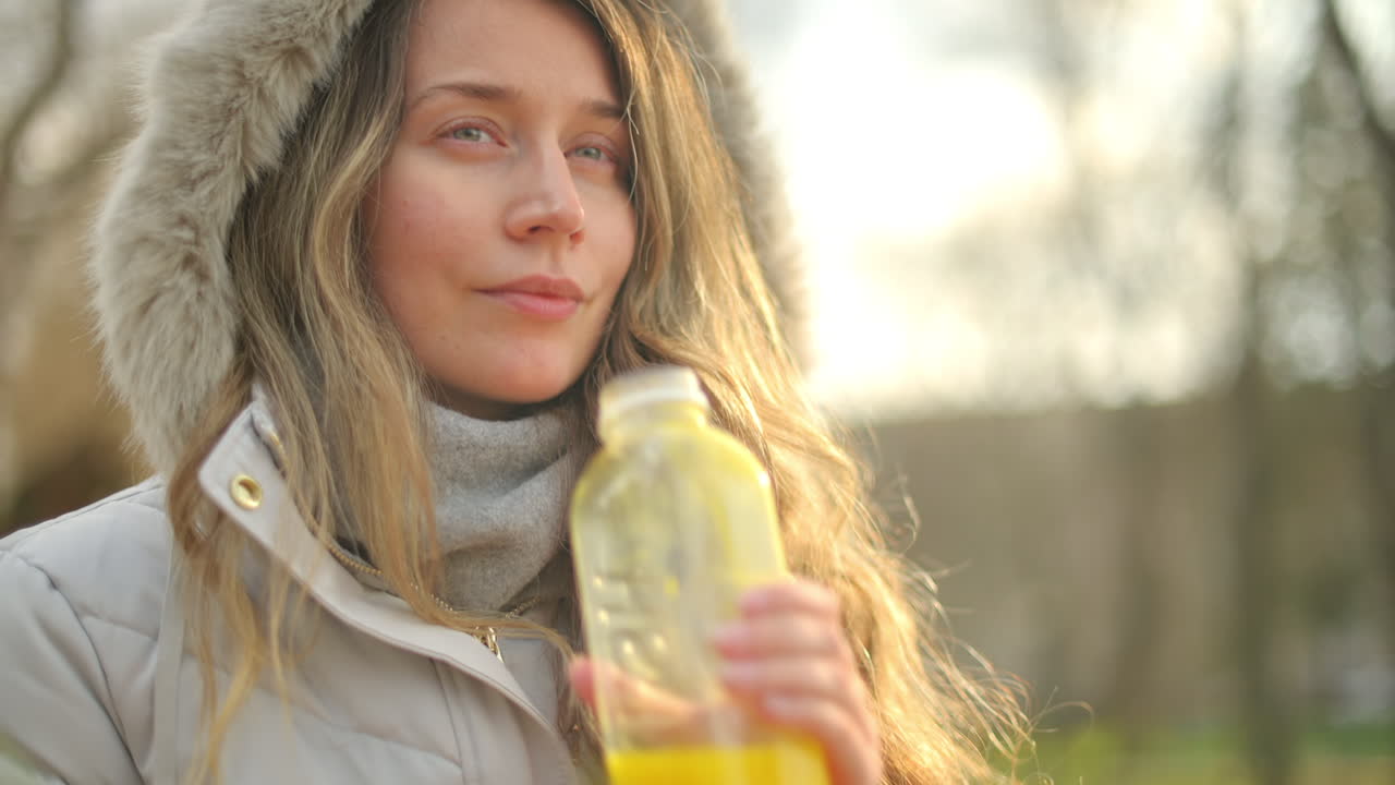 Woman in a puffer jacket drinking orange juice in a park