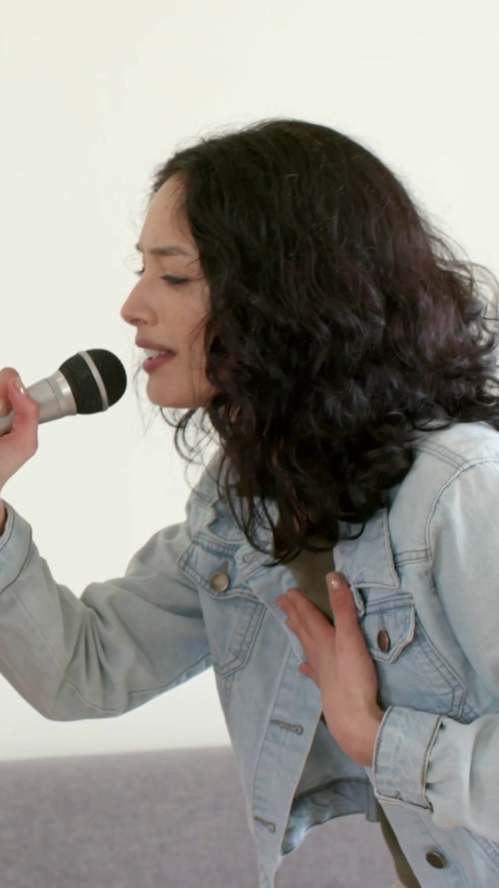Vertical video: Singing woman taking offered mic, hand on chest, rehearsing at home in denim jacket