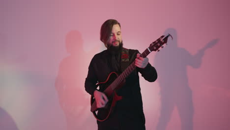 a guitarist in a black outfit stands and plays an electric guitar, moving slowly against a colorful background. the shadows on the wall create a dynamic visual effect