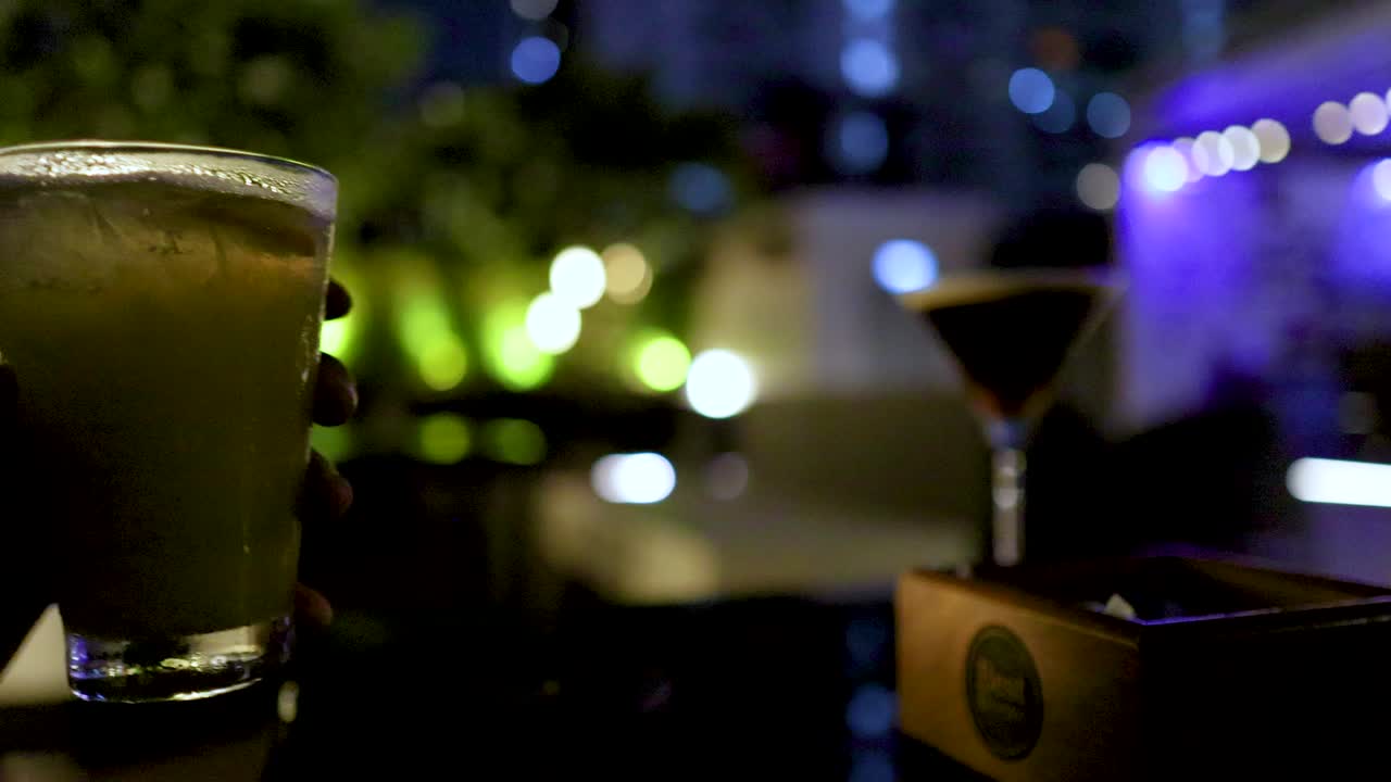 Hand picks up cocktail glass in dimly lit rooftop bar with colorful bokeh city lights