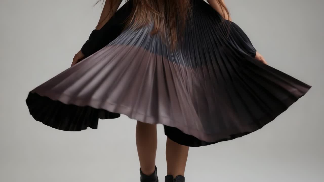 Modella asiatica che tiene i bordi di una gonna nera plissettata, che volteggia in uno studio grigio, enfatizzando le pieghe