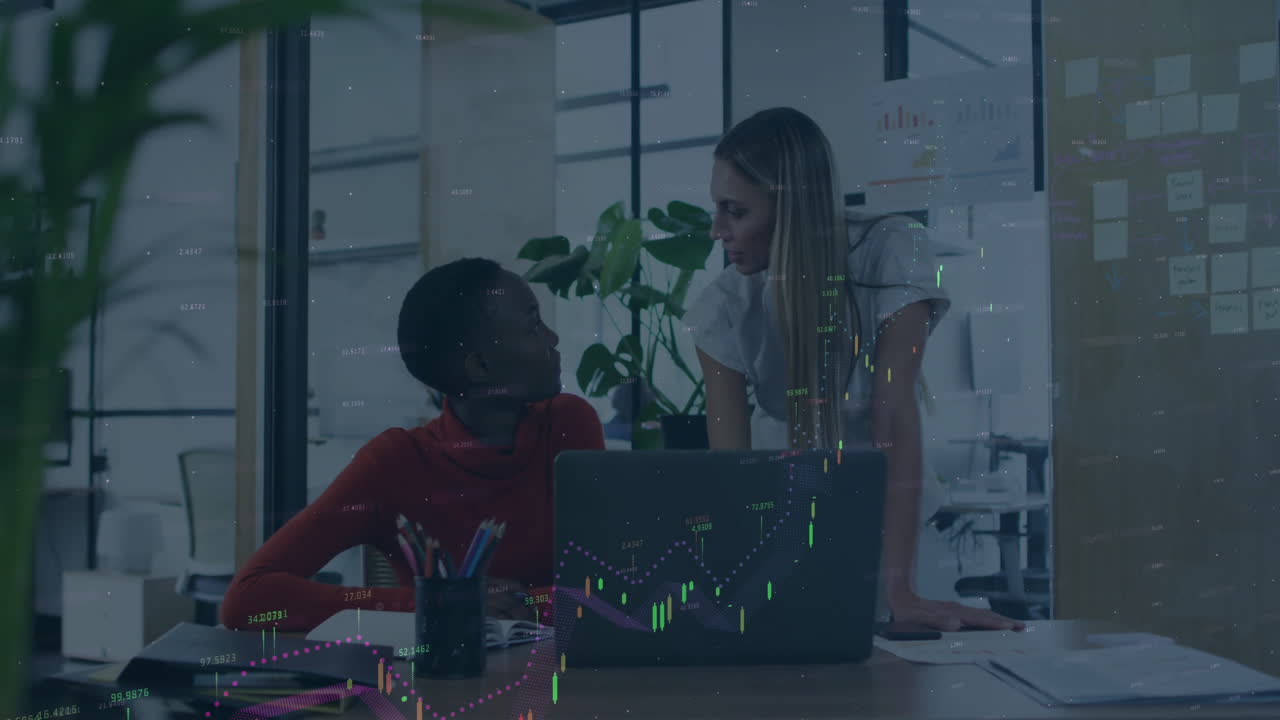 Two women collaborating at office desk with financial data animation overlay