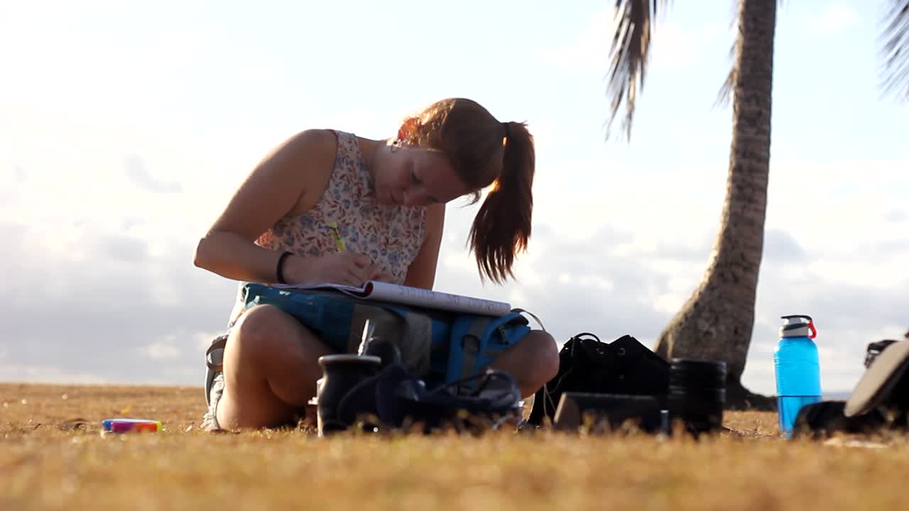 Close up shot of young woman resting on dried grass field at beach and writing letter during sunset