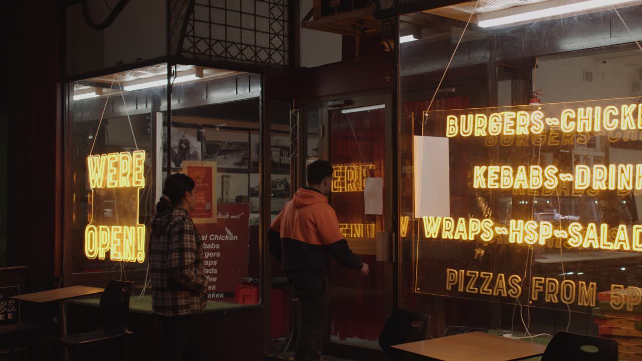 Two people enter a brightly lit fast food shop at night, neon signs glowing